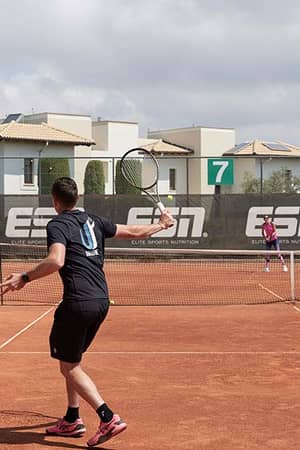 Tennis match on a clay court at Aphrodite Hills Resort with two players in action, surrounded by Mediterranean-style buildings and Elite Sports Nutrition branding.