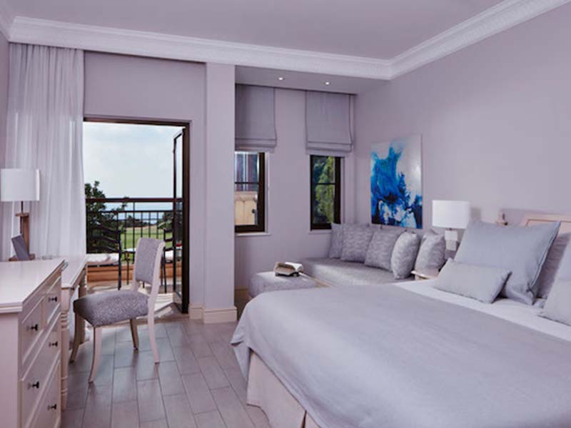 Deluxe Double Room at Aphrodite Hills with grey bedding, seating area, balcony view of greenery and sea.