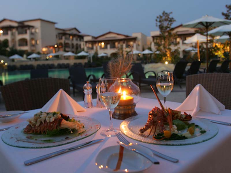 A beautifully set dining table for two with gourmet meals and a candle, overlooking a luxurious resort pool at Aphrodite Hills.