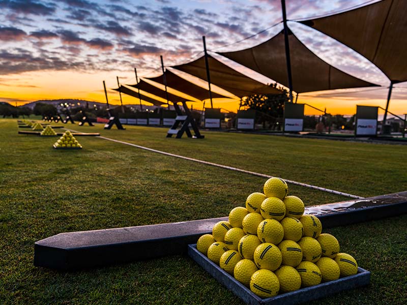 Sunset view of Aphrodite Hills driving range with stacks of yellow golf balls and canopy shades.