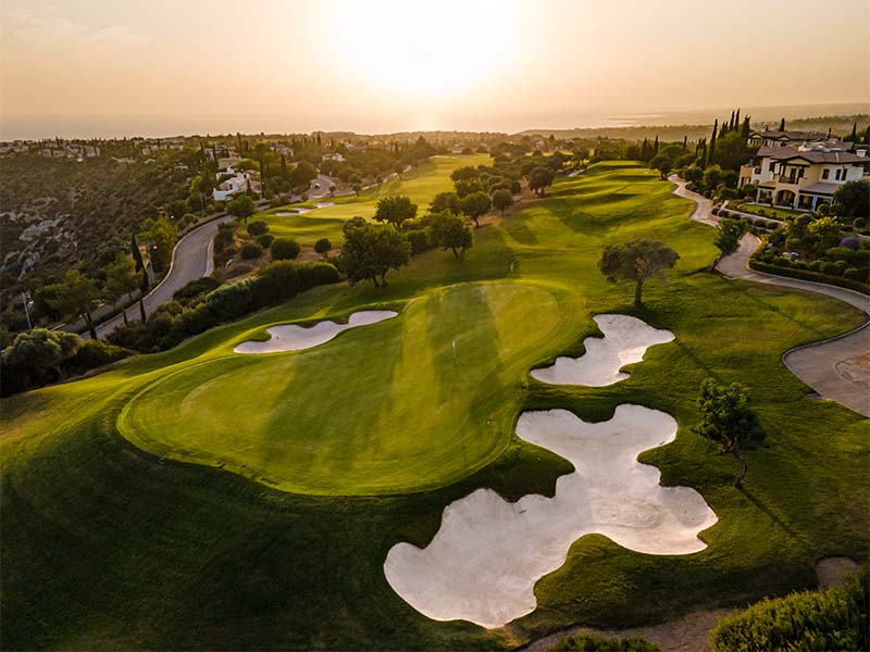 Aerial view of Aphrodite Hills Golf Course in Cyprus with green fairways, bunkers, sunset, and scenic surrounding landscapes.