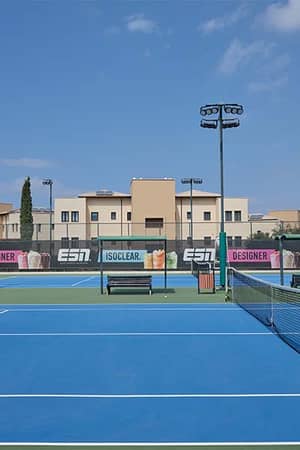 Hard tennis court at Aphrodite Hills with blue surface and advertisements, set against a modern building and partly cloudy sky.