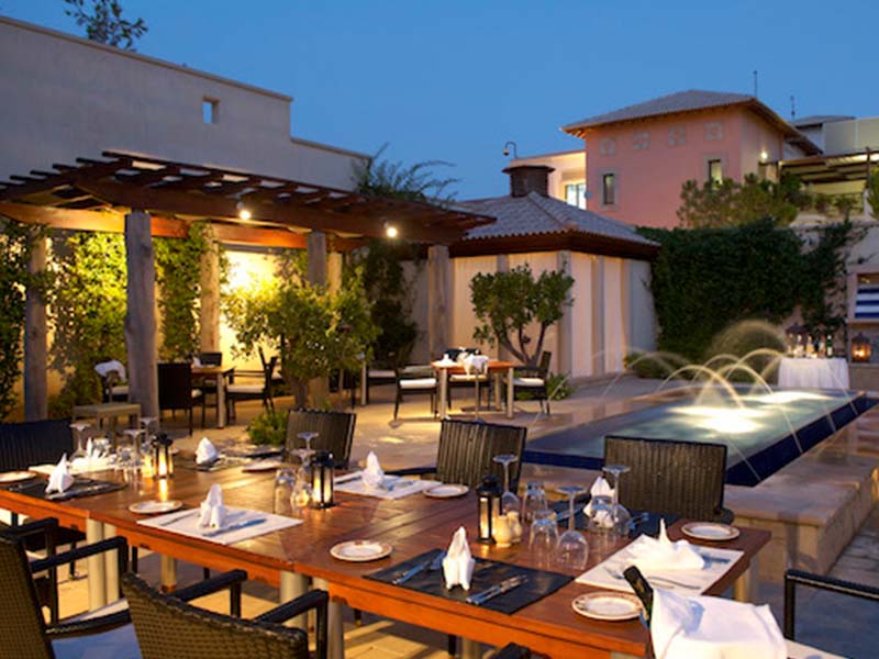 Outdoor dining area of Aphrodite Hills Resort's main restaurant in the evening, featuring elegantly set tables and a lit water feature.