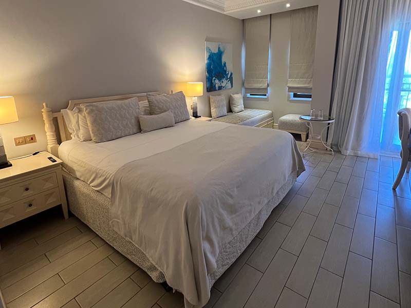 Hotel room at Aphrodite Hills with a double bed, white bedding, bedside lamps, decorative wall art, and large windows with grey curtains.
