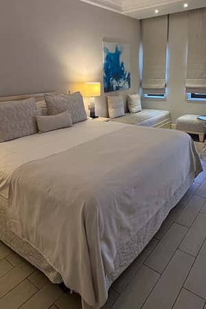 Hotel room at Aphrodite Hills with a double bed, white bedding, bedside lamps, decorative wall art, and large windows with grey curtains.