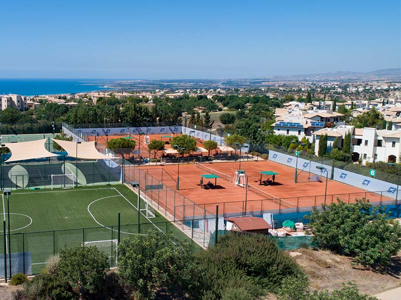 Aerial view of clay tennis courts and a football pitch at Aphrodite Hills Resort, surrounded by villas and overlooking the Mediterranean Sea.