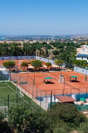 Aerial view of clay tennis courts and a football pitch at Aphrodite Hills Resort, surrounded by villas and overlooking the Mediterranean Sea.