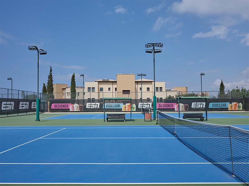 Tennis court at Aphrodite Hills with blue and green surfaces, adverts around, and modern villas in the background.