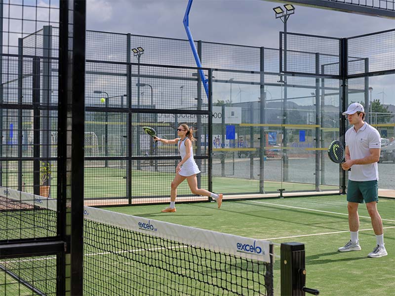 Two players on a padel court at Aphrodite Hills Resort, a woman serving and a man preparing to play under cloudy skies.