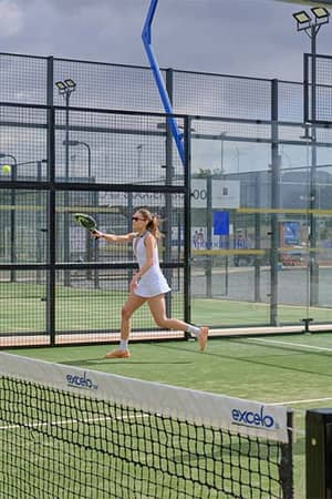 Two players on a padel court at Aphrodite Hills Resort, a woman serving and a man preparing to play under cloudy skies.