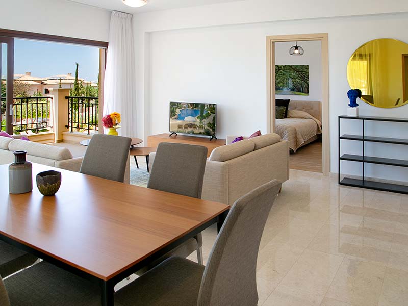 Open-plan living and dining area in an Aphrodite Hills villa, featuring modern furnishings, a dining table, sofa, TV, bedroom access, and balcony with views.