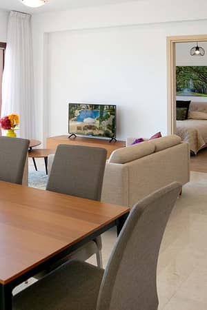 Open-plan living and dining area in an Aphrodite Hills villa, featuring modern furnishings, a dining table, sofa, TV, bedroom access, and balcony with views.