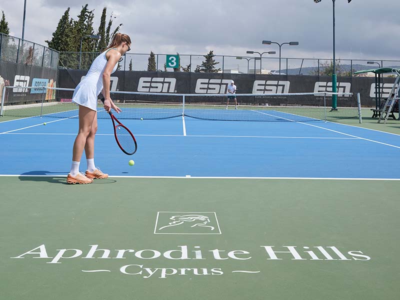 Woman getting ready to serve on a tennis court at Aphrodite Hills Resort, Cyprus, with the Aphrodite Hills logo on the ground.