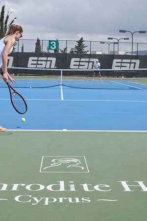 Woman getting ready to serve on a tennis court at Aphrodite Hills Resort, Cyprus, with the Aphrodite Hills logo on the ground.