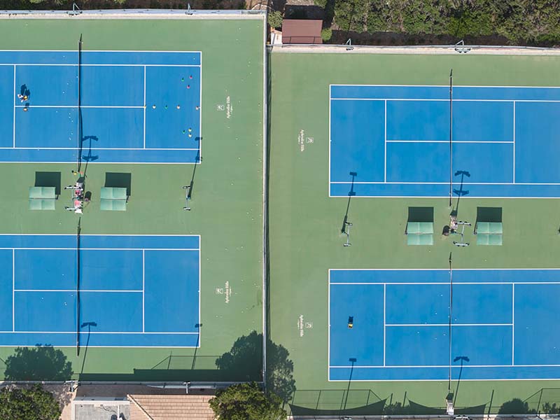 Aerial view of tennis courts at Aphrodite Hills with blue playing surfaces and green surroundings, showcasing seating and nets.