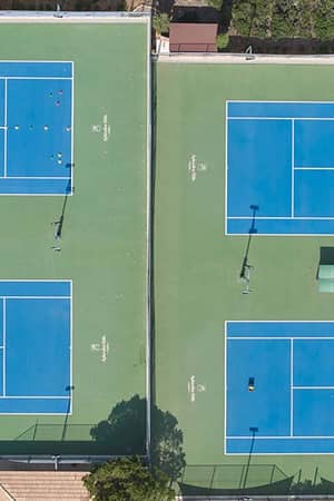 Aerial view of tennis courts at Aphrodite Hills with blue playing surfaces and green surroundings, showcasing seating and nets.
