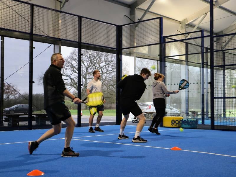 Four people engaged in an indoor padel tennis coaching session, playing on a blue court with padel racquets.
