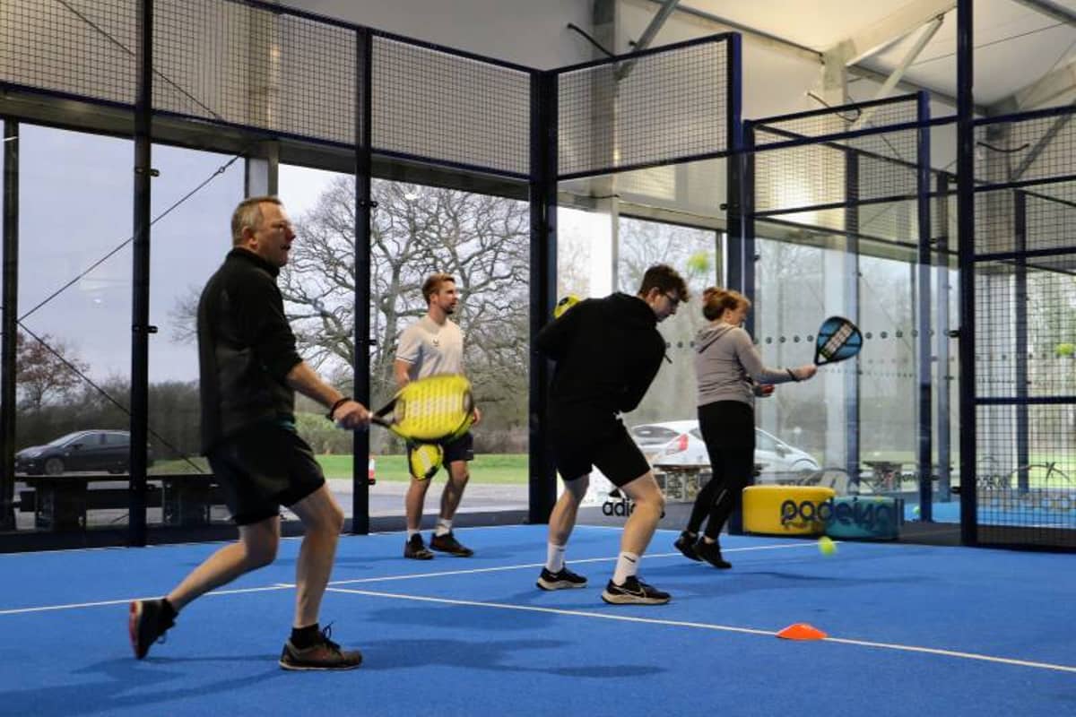 Four people engaged in an indoor padel tennis coaching session, playing on a blue court with padel racquets.