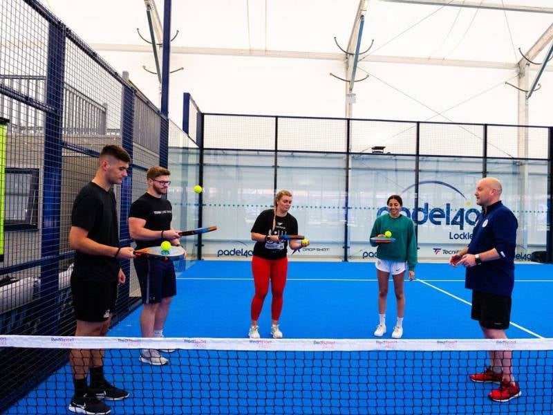 Padel tennis coaching session at BD Swindon Gallery with players and a coach on a blue court.