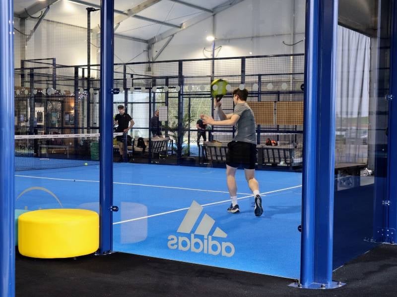 Indoor padel court with players in action, featuring blue floor and glass walls, with visible adidas logo.