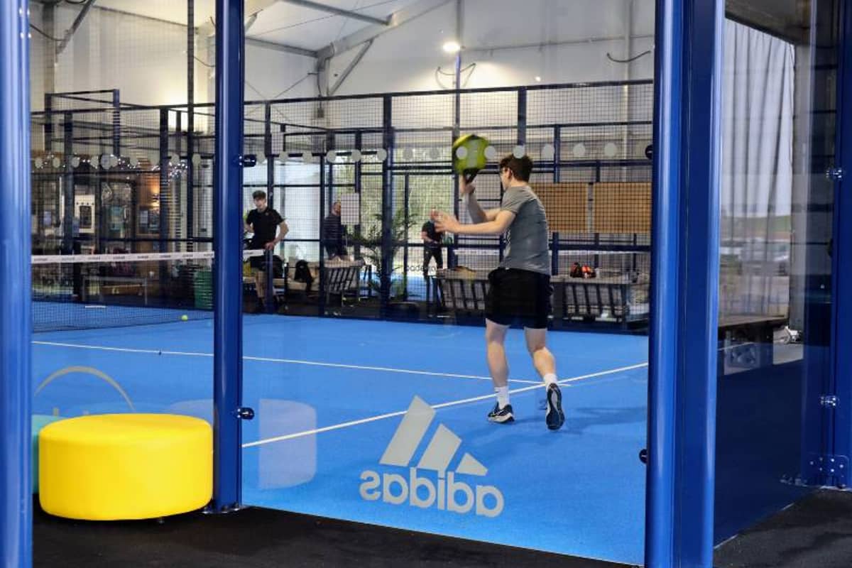 Indoor padel court with players in action, featuring blue floor and glass walls, with visible adidas logo.