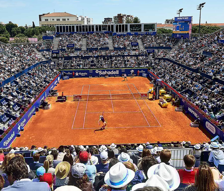Panoramic view of a tennis match on a clay court at the Barcelona Open with a full audience and a clear blue sky.