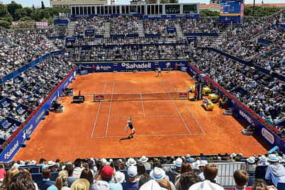 Panoramic view of a tennis match on a clay court at the Barcelona Open with a full audience and a clear blue sky.