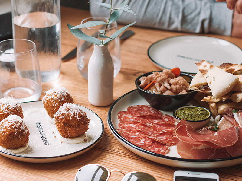 Assortment of Spanish tapas in Barcelona, featuring arancini balls, charcuterie platter with sliced meats, and pesto with pickled vegetables on a wooden table.