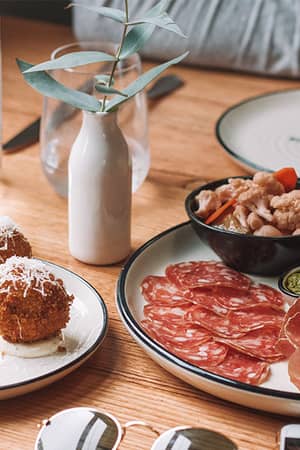 Assortment of Spanish tapas in Barcelona, featuring arancini balls, charcuterie platter with sliced meats, and pesto with pickled vegetables on a wooden table.