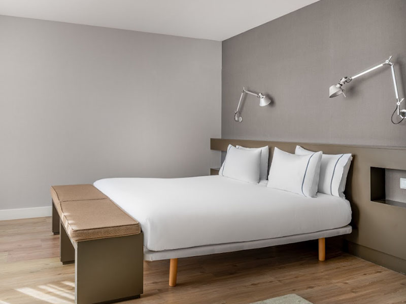 Minimalist hotel room in Barcelona with a double bed, white linens, wall-mounted lamps, and a sleek bench at the bed's foot.