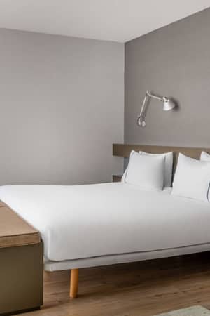 Minimalist hotel room in Barcelona with a double bed, white linens, wall-mounted lamps, and a sleek bench at the bed's foot.