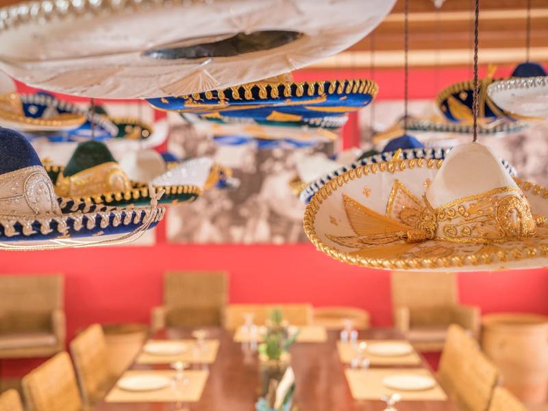 Dining area with colourful sombreros hanging from the ceiling and a large wooden table set for a meal at a beach club.