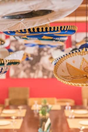 Dining area with colourful sombreros hanging from the ceiling and a large wooden table set for a meal at a beach club.