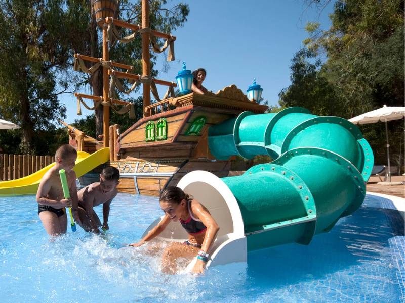 Children playing on a water slide at a beach club, with a pirate ship backdrop and a girl sliding into a shallow pool.