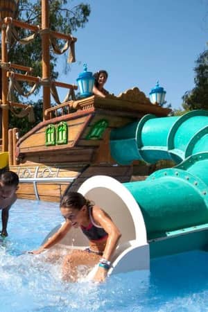 Children playing on a water slide at a beach club, with a pirate ship backdrop and a girl sliding into a shallow pool.