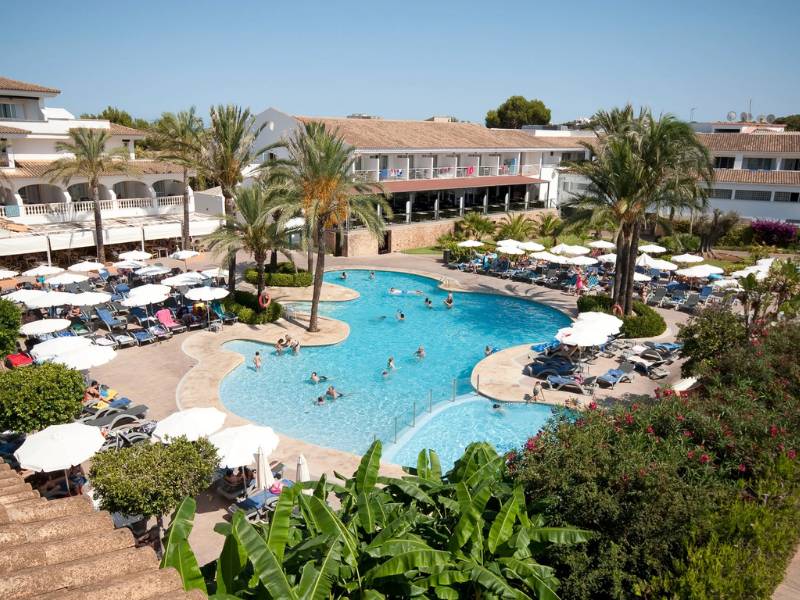 Outdoor swimming pool area at a beach club with loungers, parasols, and palm trees, set against a Mediterranean-style building.