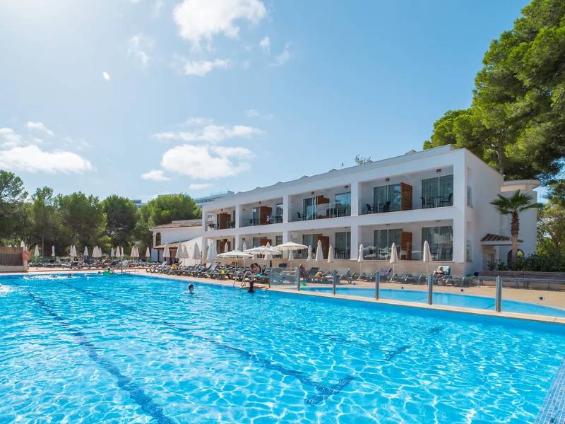 Outdoor pool area at a beach club with a clear blue pool, sun loungers, parasols, and a two-storey building surrounded by trees.