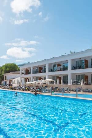 Outdoor pool area at a beach club with a clear blue pool, sun loungers, parasols, and a two-storey building surrounded by trees.