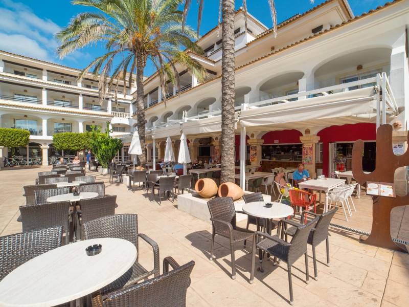 Outdoor dining area at a beach club with tables, chairs, white parasols, and a Mediterranean-style building in the background.