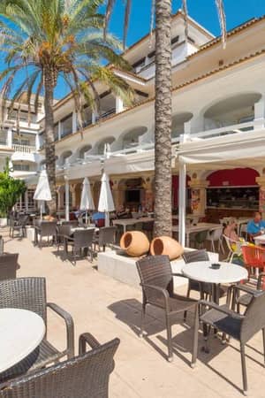 Outdoor dining area at a beach club with tables, chairs, white parasols, and a Mediterranean-style building in the background.