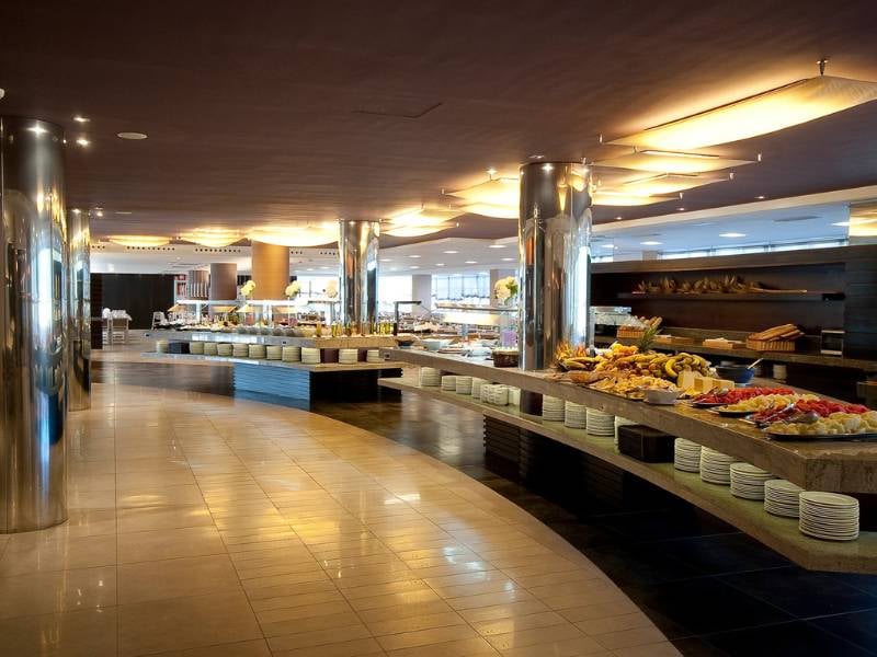 Luxurious beach club buffet with curved counters and an assortment of fruits, pastries, and gourmet dishes under ambient lighting.