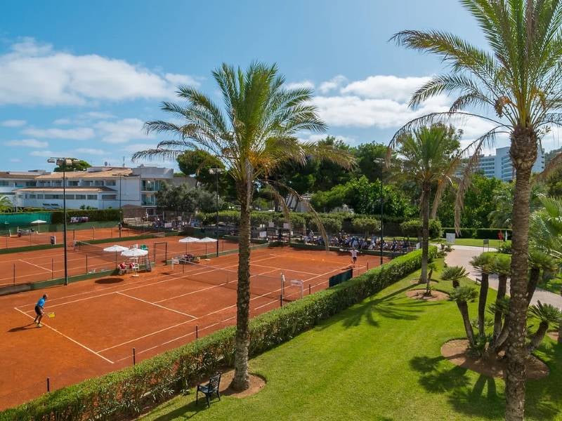 Tennis courts at a beach club surrounded by greenery and palm trees, with players in action against a backdrop of club facilities and clear skies.