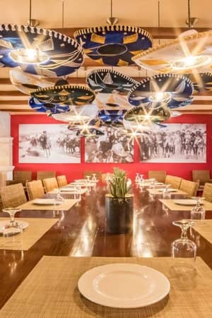 Elegant dining room at a beach club with a wooden table, woven placemats, white plates, and decorative umbrella lights, set against coral walls with a beach scene mural.
