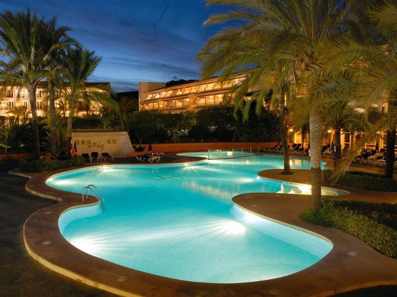 Night view of a beach club swimming pool surrounded by palm trees, with a hotel building illuminated in the background.