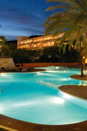 Night view of a beach club swimming pool surrounded by palm trees, with a hotel building illuminated in the background.