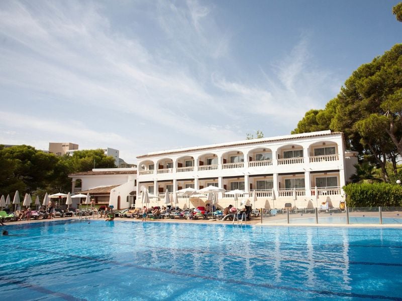 Outdoor pool at a beach club, with a Mediterranean-style building, surrounded by sunbathers and pine trees.