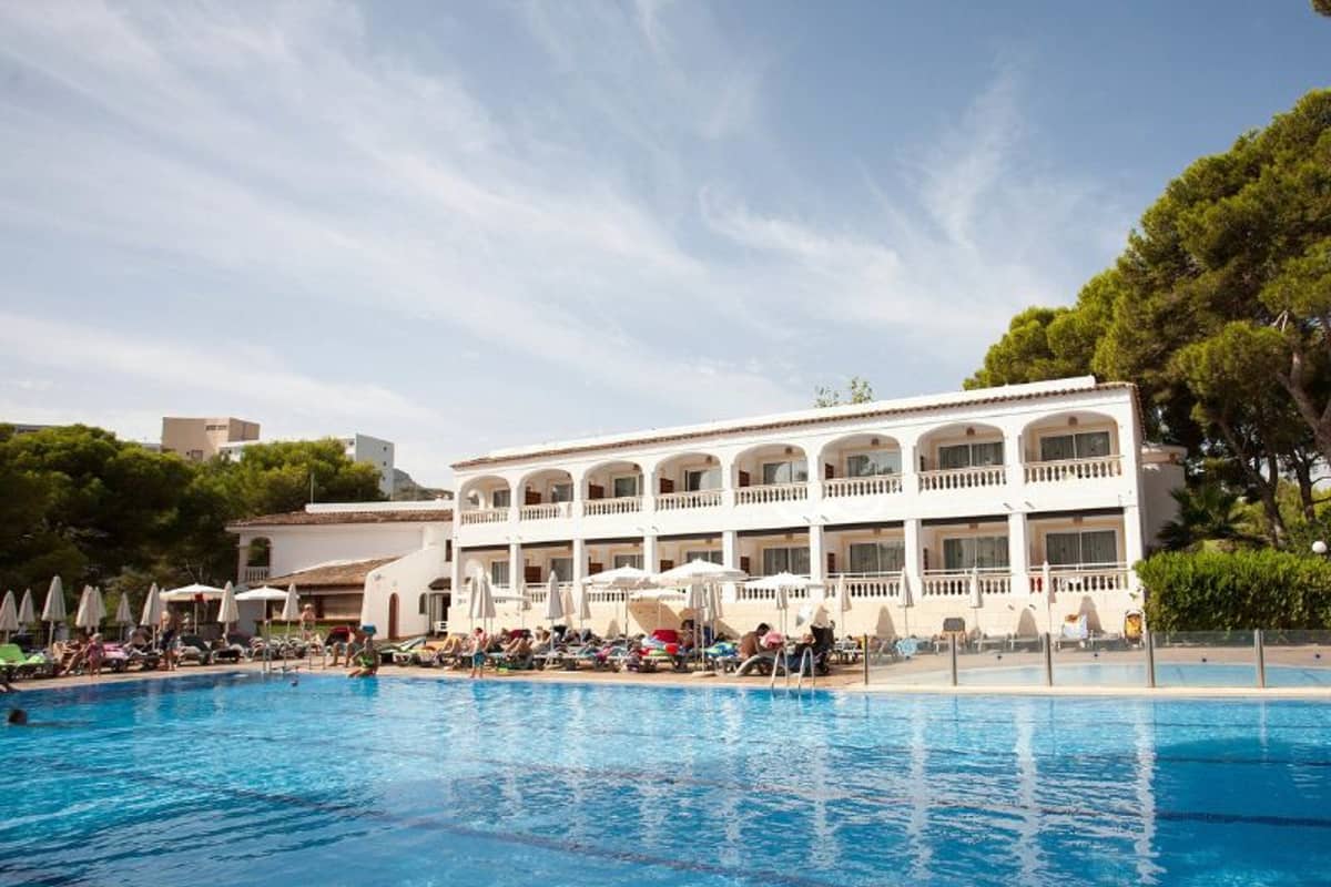 Outdoor pool at a beach club, with a Mediterranean-style building, surrounded by sunbathers and pine trees.