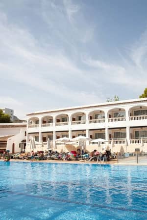 Outdoor pool at a beach club, with a Mediterranean-style building, surrounded by sunbathers and pine trees.