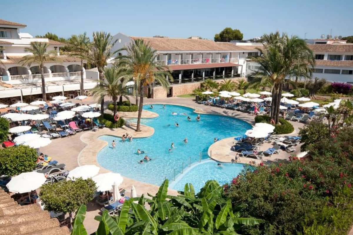 Aerial view of Beach Club Es Talaial with a large swimming pool, sun loungers, and tropical palm trees.