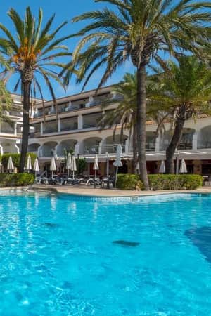 Swimming pool at a beach club with clear blue water, surrounded by sun loungers and palm trees, hotel building in the background.